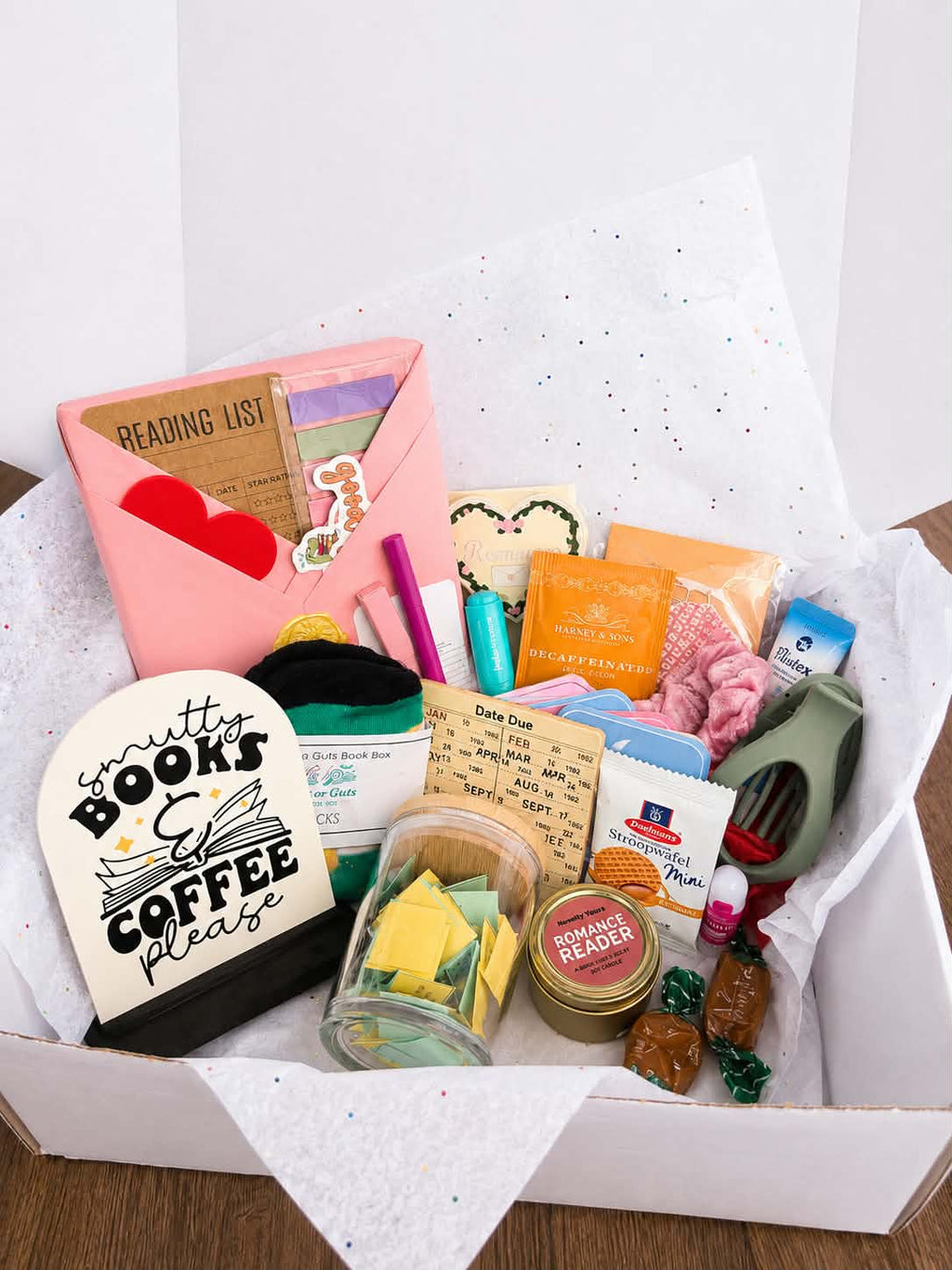 PAST BOX -

A curated book and coffee gift box filled with various themed items including a pink reading list envelope, a 'Smartly Books & Coffee Please' sign, a glass jar of tea bags, a 'Fiction Reader' tin, and colorful stationery, all arranged in white tissue paper inside a white box.